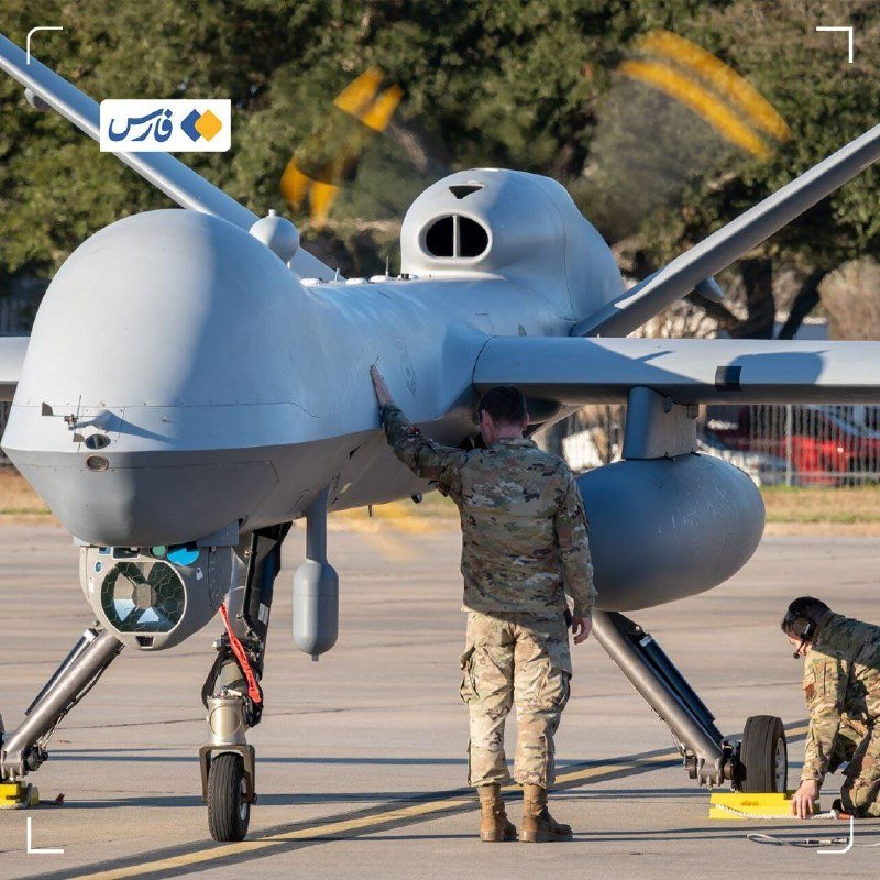 Two uniformed military personnel conduct maintenance on a gray unmanned aerial vehicle parked on a tarmac, one wiping its wing and another crouched near the rear wheel.