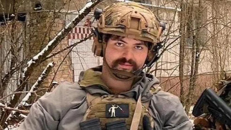 A man with a mustache wearing a camouflage tactical helmet and body armor stands outdoors near bare trees, looking at the camera.