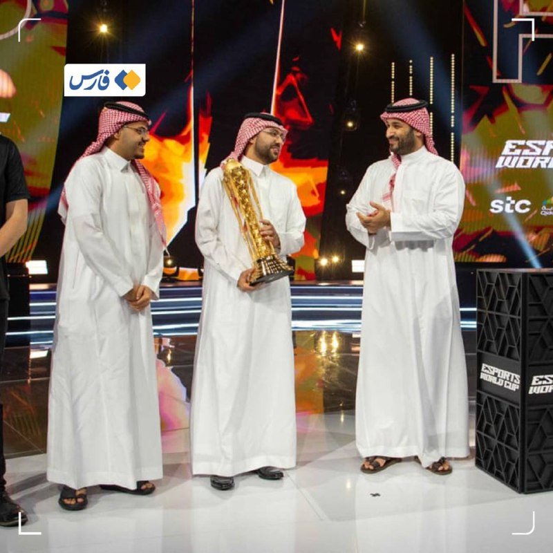Three men in traditional white thobes and red-checkered keffiyehs stand on a brightly lit stage, with the central figure holding a large golden trophy during an awards ceremony.