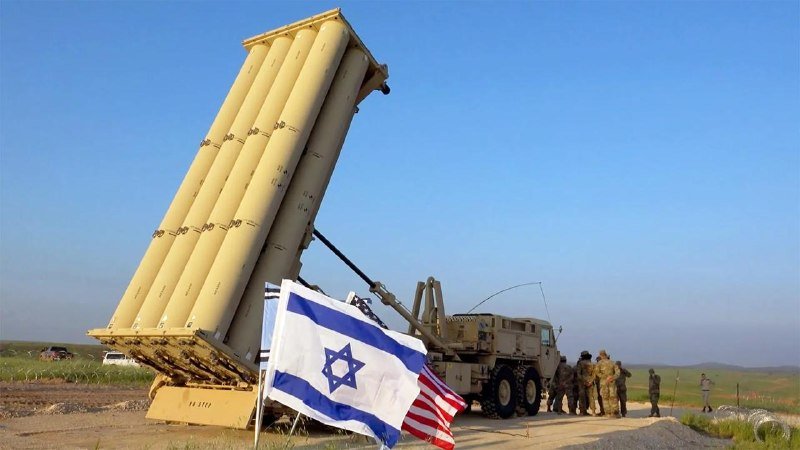 A missile launcher system is elevated beside an Israeli flag, with military personnel standing nearby on a paved road in an open landscape.
