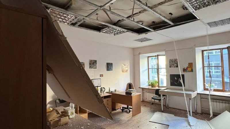 A damaged office interior with a collapsed ceiling, scattered debris, overturned furniture, and an open door amidst the destruction.