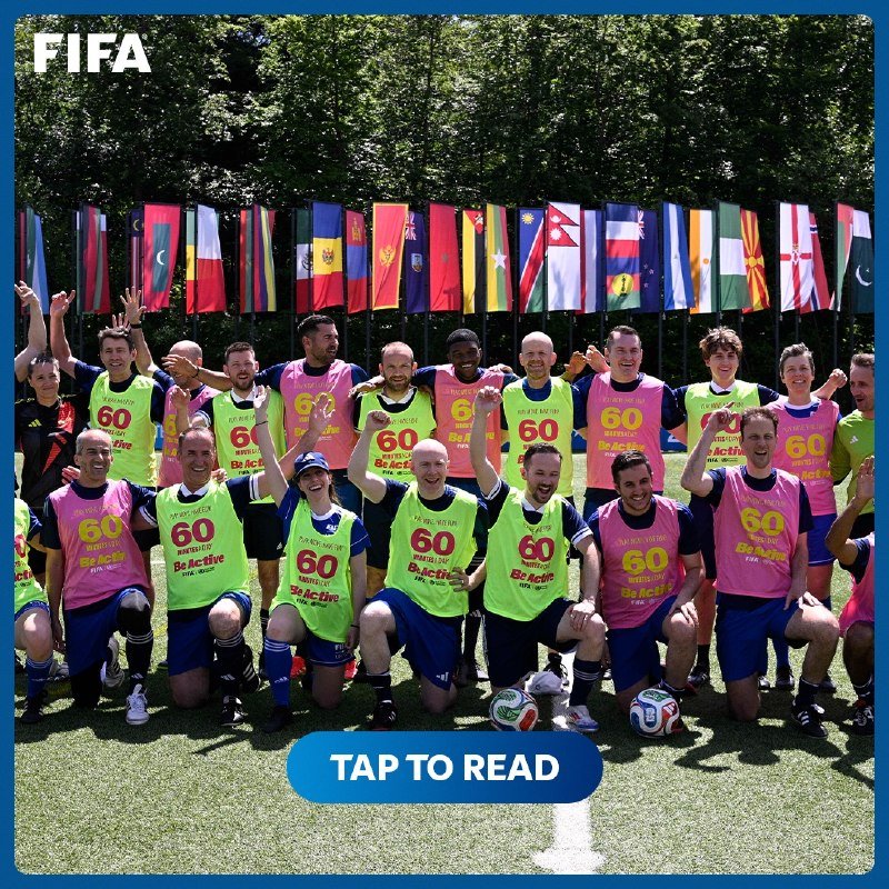 A group of people wearing yellow and pink bibs marked "60 Be Active" pose together on grass in front of a row of international flags, with a FIFA logo visible.