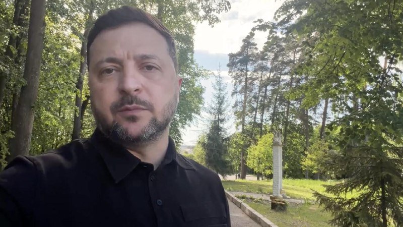 A bearded man in a black shirt takes a selfie outdoors among tall trees, with a stone monument visible in the background.