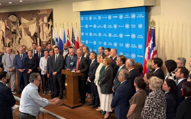 A man in a red tie speaks at a wooden podium flanked by officials and international flags before a United Nations Security Council backdrop.
