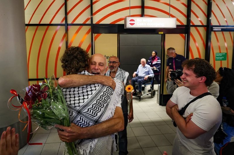 Two men wearing keffiyehs embrace while holding flowers at an airport boarding gate, as onlookers smile nearby.