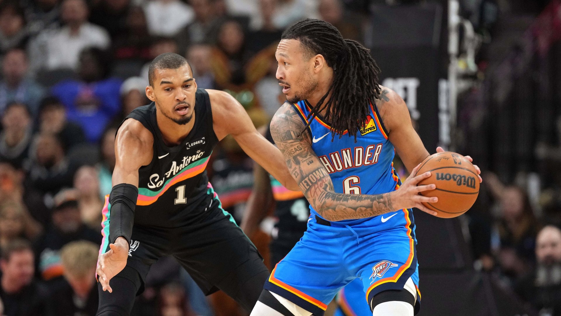 An Oklahoma City Thunder player in a blue #6 jersey holds a basketball while being defended by a San Antonio Spurs player in a black #1 jersey during an NBA game.