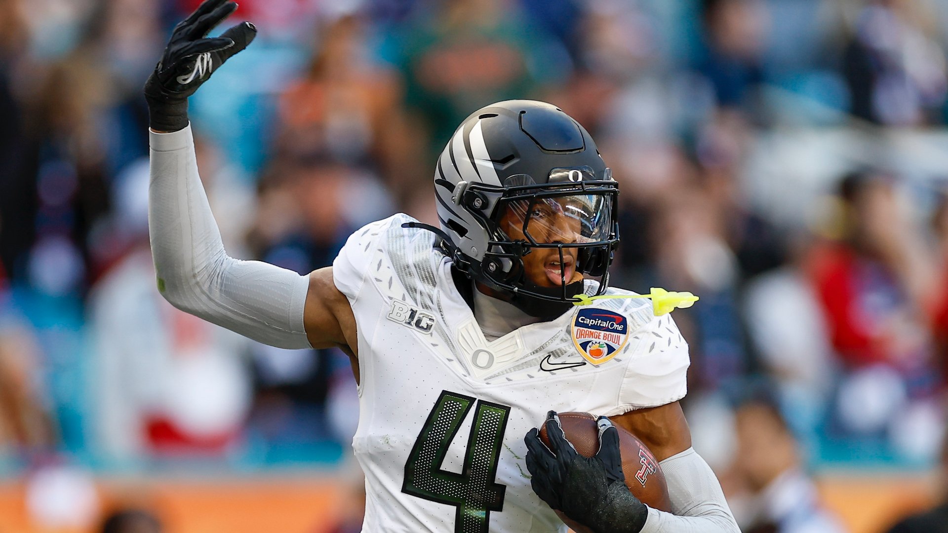 A football player wearing a white jersey with the number 4, a silver helmet, and a Capital One Orange Bowl patch runs with the ball while raising one arm, with a blurred crowd in the background.