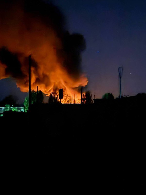 A large fire burns intensely at night, sending thick orange smoke into the sky above silhouetted trees, vehicles, and utility structures.