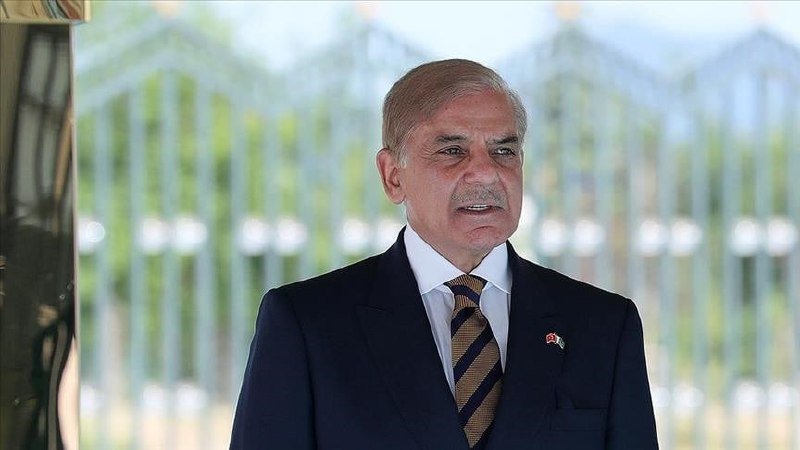 A gray-haired man in a dark suit and striped tie stands outdoors in front of a metal gate, his mouth open as if speaking.