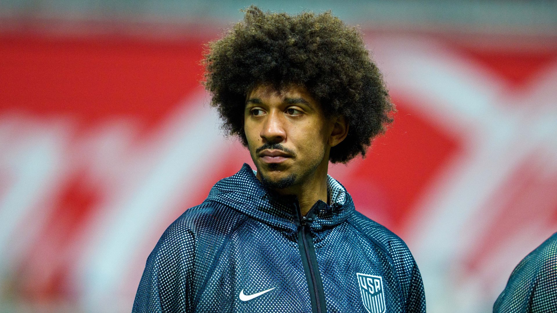 A man with a large afro stands before a red and white background, wearing a dark USA soccer training jacket.