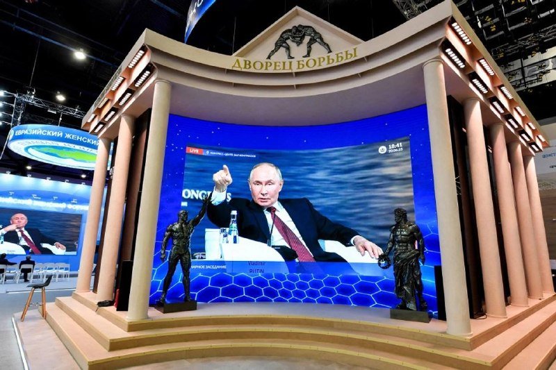 A man in a suit gestures while speaking on a large screen displayed within a classical-style stage featuring columns and the Russian text "ДВОРЕЦ БОРЬБЫ."
