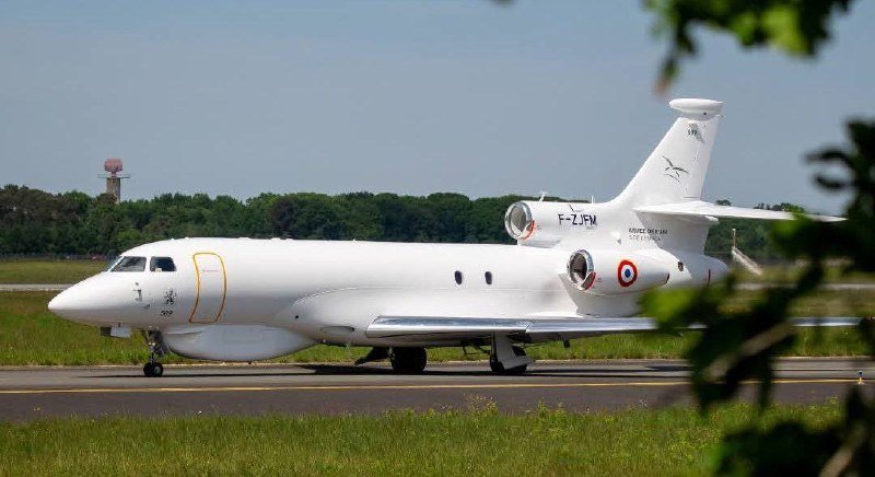 A white tri-jet aircraft with French military roundels and registration "F-ZJPM" taxis on a runway, with trees and a tower visible in the background.