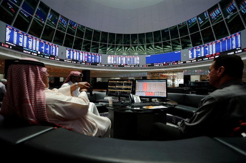 Three men, two in traditional Arab headdresses and one in a suit, sit at desks beneath large curved digital stock ticker displays inside a domed trading floor.