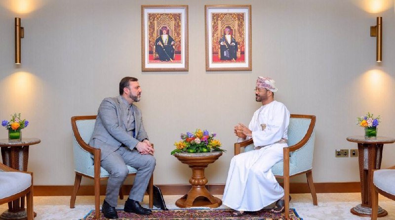 Two men sit facing each other in armchairs during a meeting, one in a gray suit and the other in traditional white attire and turban, with floral arrangements and two framed portraits on the wall behind them.