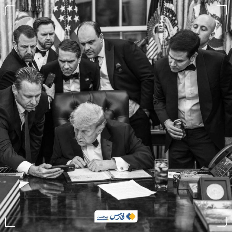 Black-and-white photo of seven men in tuxedos gathered around an older seated man reviewing documents at a desk, with American flags visible in the background.