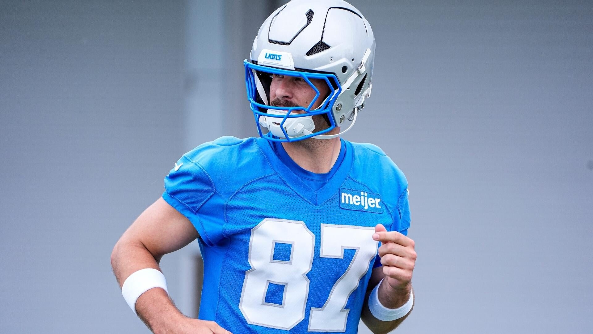 A football player wearing a blue Detroit Lions jersey numbered 87 and a silver helmet with blue facemask runs against a gray background.