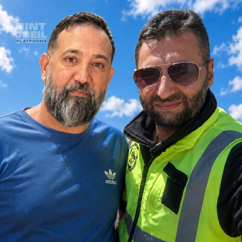 Two bearded men pose for a close-up selfie outdoors against a blue sky; one wears a blue Adidas t-shirt, the other wears sunglasses and a high-visibility yellow safety vest.