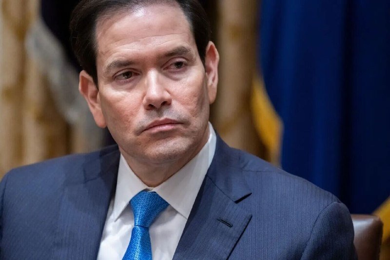 A dark-haired man in a navy suit and blue tie sits with a serious expression before a blue flag and gold curtains.
