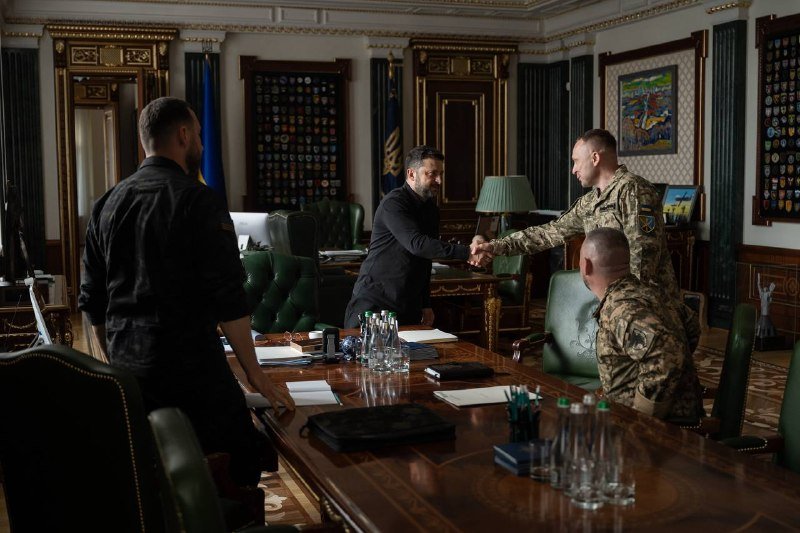 A man in dark clothing shakes hands with a man in military camouflage uniform inside an ornately decorated room with green leather chairs and a large wooden conference table.