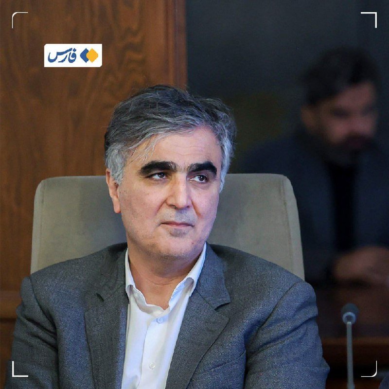 A gray-haired man in a gray blazer and white shirt sits in a chair, looking off to the side, with another person visible in the blurred background.