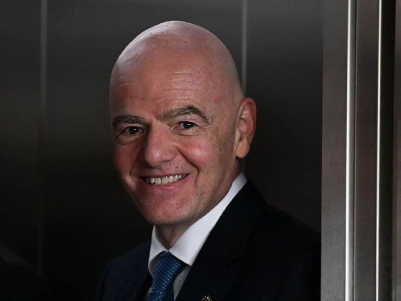 A bald man in a dark suit, white shirt, and blue tie smiles while standing against a dark background.
