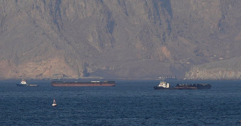 Three large cargo ships float on calm blue water near a small buoy, set against towering rocky mountains in the background.