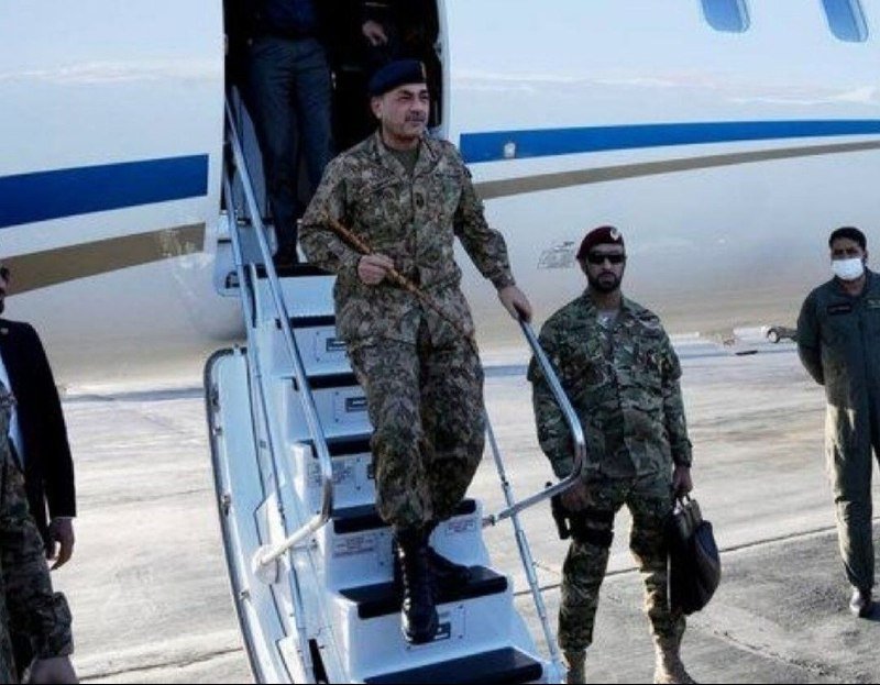 A military officer in camouflage uniform descends the stairs of a white aircraft on a tarmac, accompanied by another uniformed soldier wearing a maroon beret.