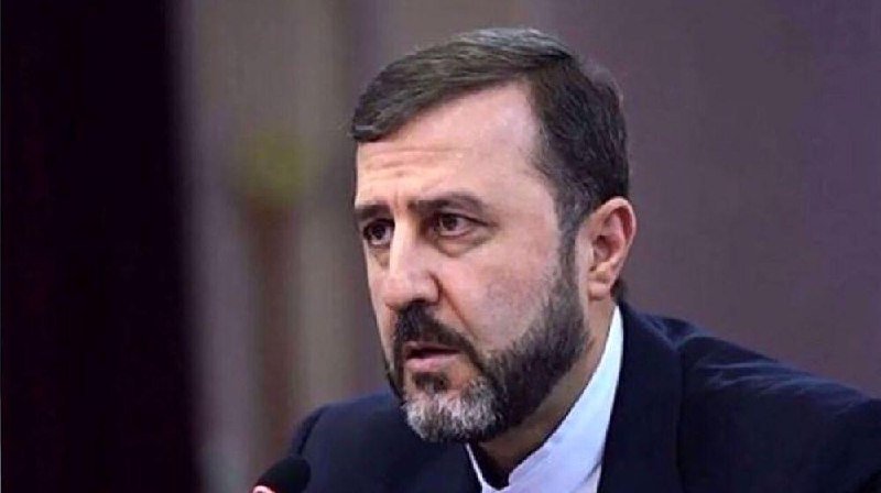 A bearded man in a dark suit and white shirt speaks into a microphone against a blurred background.