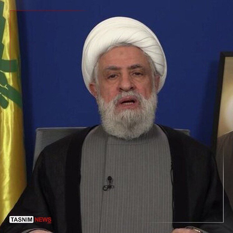 A bearded man wearing a white turban and dark clerical robes speaks into a lapel microphone, with a yellow Hezbollah flag visible beside him against a blue backdrop.