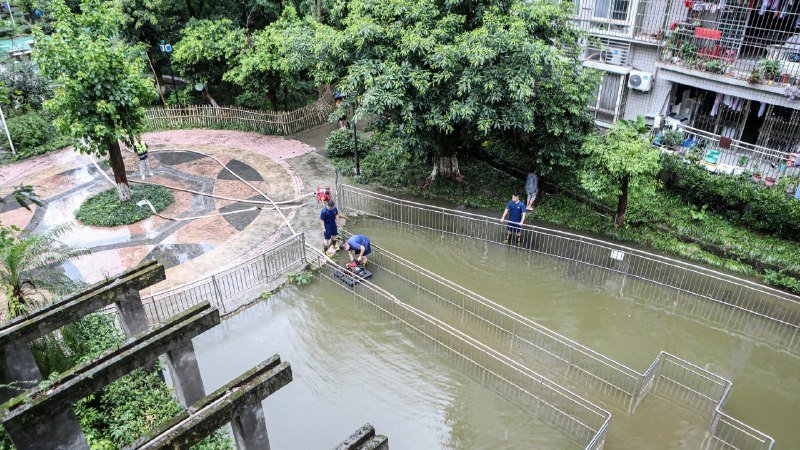 Several people work around a flooded, fenced pathway next to a residential building and tree-lined park area.