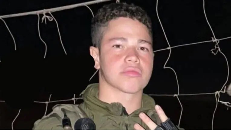 A young man with short curly hair wearing an olive-green hooded jacket stands in front of a barbed-wire fence at night, appearing to speak.