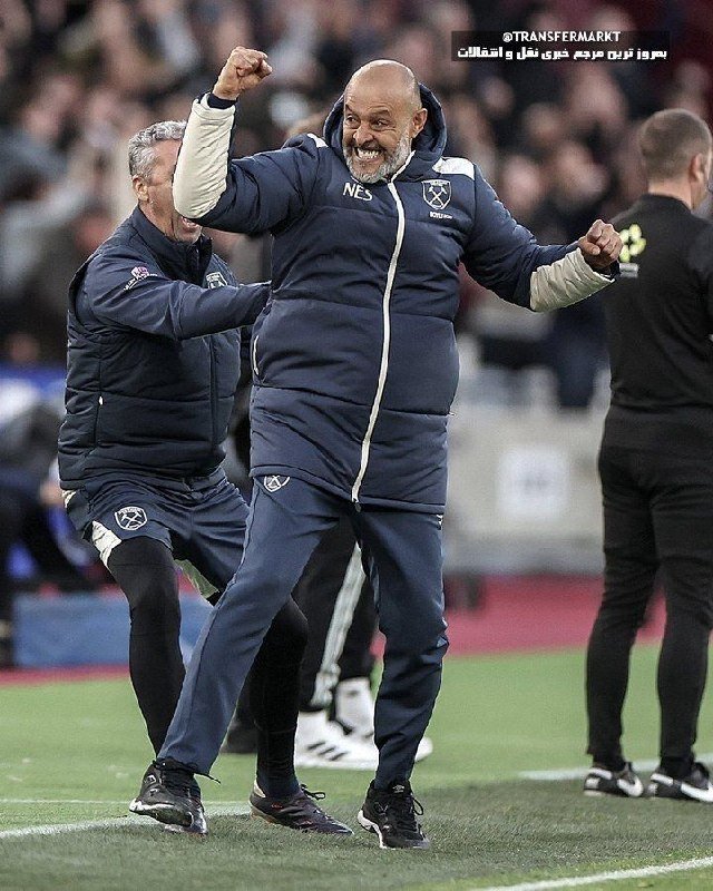 A bald, bearded man in a navy tracksuit jacket with "NES" lettering pumps his fist and smiles on a football pitch sideline, with another man in matching team gear behind him.