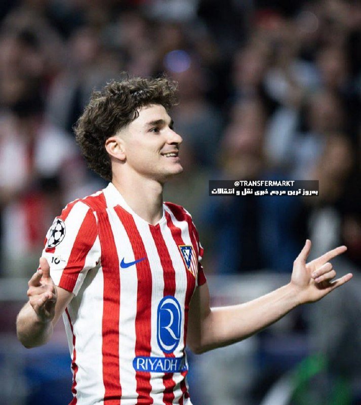 A curly-haired soccer player in a red and white striped jersey smiles and points while standing before a blurred crowd of spectators.