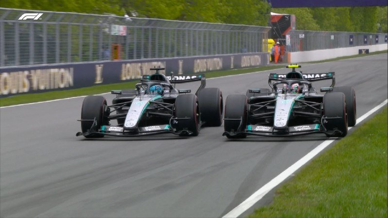 Two black Formula 1 race cars with Petronas branding drive side-by-side down a track past a "Louis Vuitton" barrier.