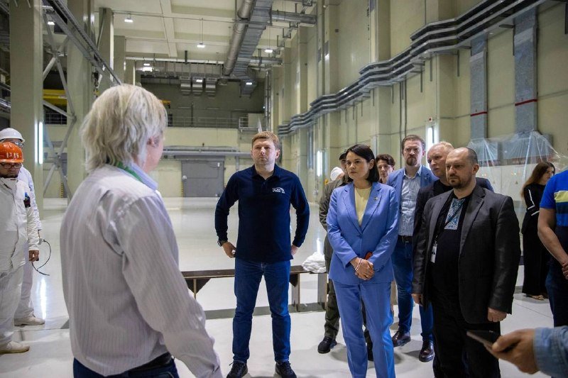 A group of people, including individuals in business attire and workers in hard hats, stand listening to a gray-haired speaker inside a large industrial facility.