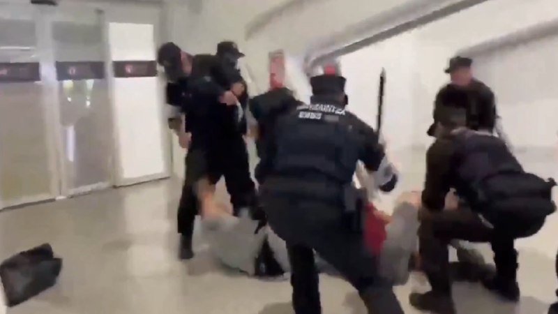 Heavily armored officers in tactical gear physically restrain multiple individuals on the floor of a bright, white-tiled corridor.