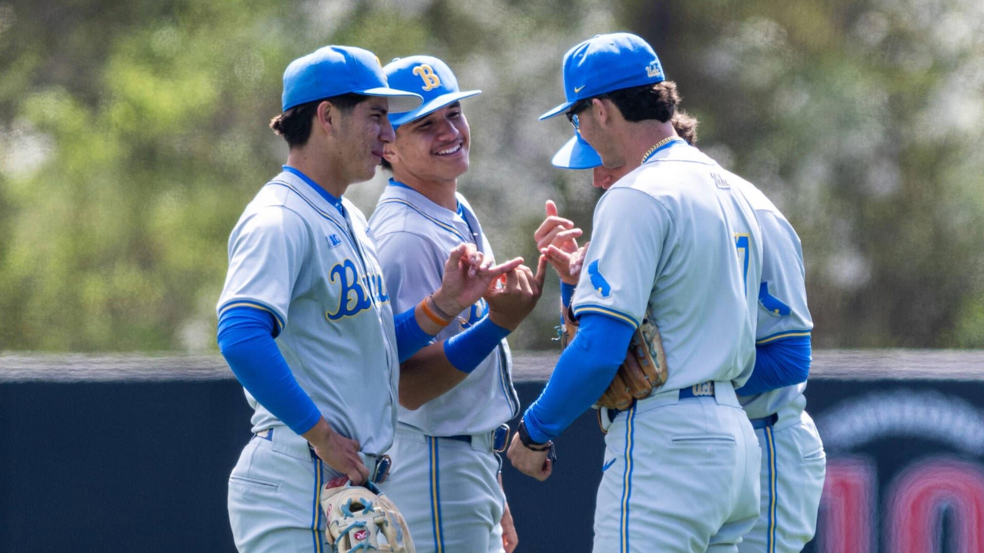 Four baseball players in gray and blue uniforms with "Bruins" lettering gather on the field, smiling and exchanging hand gestures during a game.