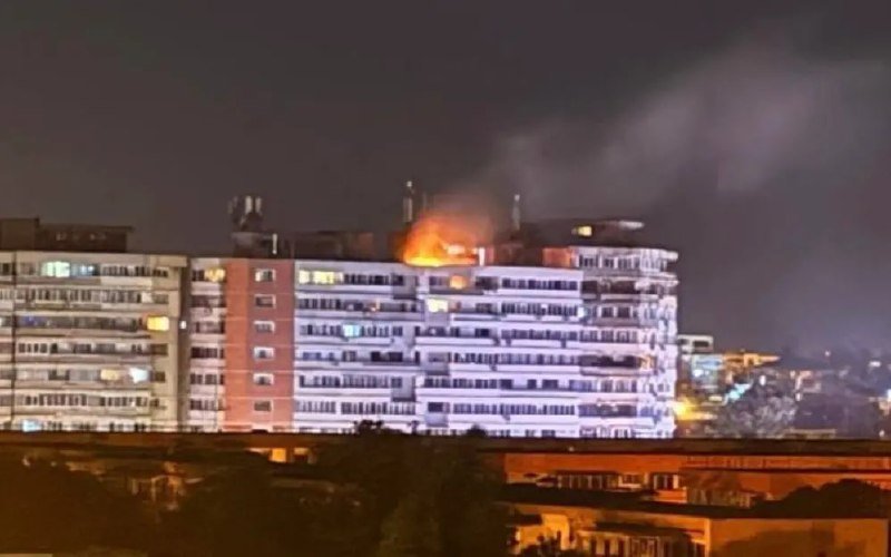 Flames and smoke rise from the rooftop of a multi-story residential building at night, with lit windows visible across the structure's facade.