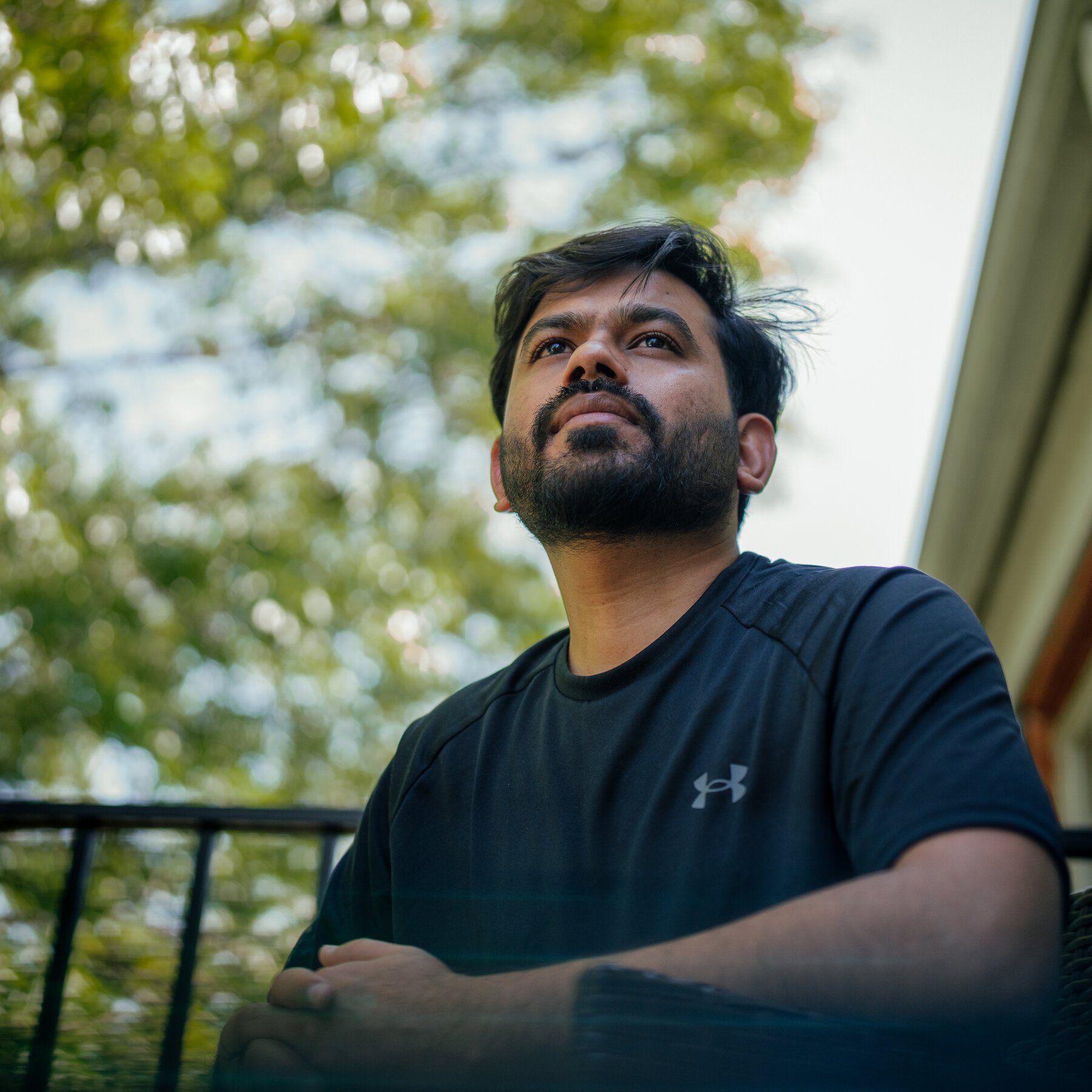 A bearded man in a dark Under Armour t-shirt sits on a balcony, gazing upward against a backdrop of green trees and sky.