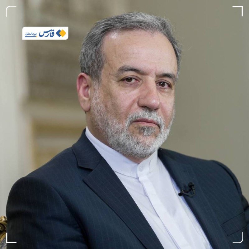 A gray-haired, bearded man in a dark suit and white shirt sits with a small lapel microphone attached, against a softly blurred indoor background.