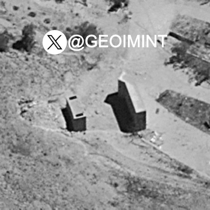 Black-and-white aerial/satellite imagery shows two dark, irregular structures casting long shadows on a barren, sandy terrain, with an "@GEOIMINT" watermark overlaid.