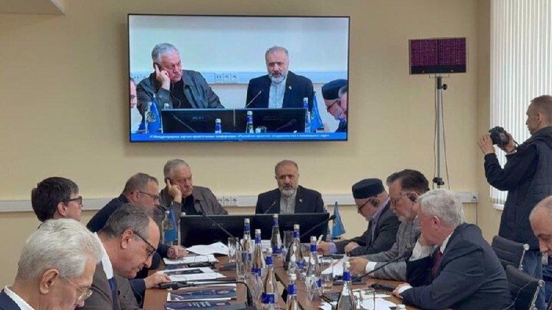 A group of men sit around a conference table covered with papers and water bottles during a meeting, with a large wall-mounted screen displaying two of the participants.