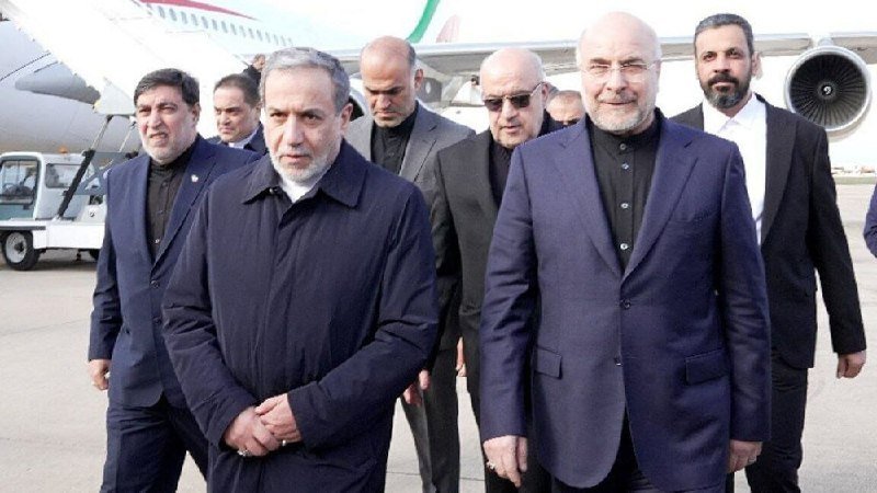 A group of men in dark suits and jackets walk together on a tarmac with a large passenger airplane visible in the background.