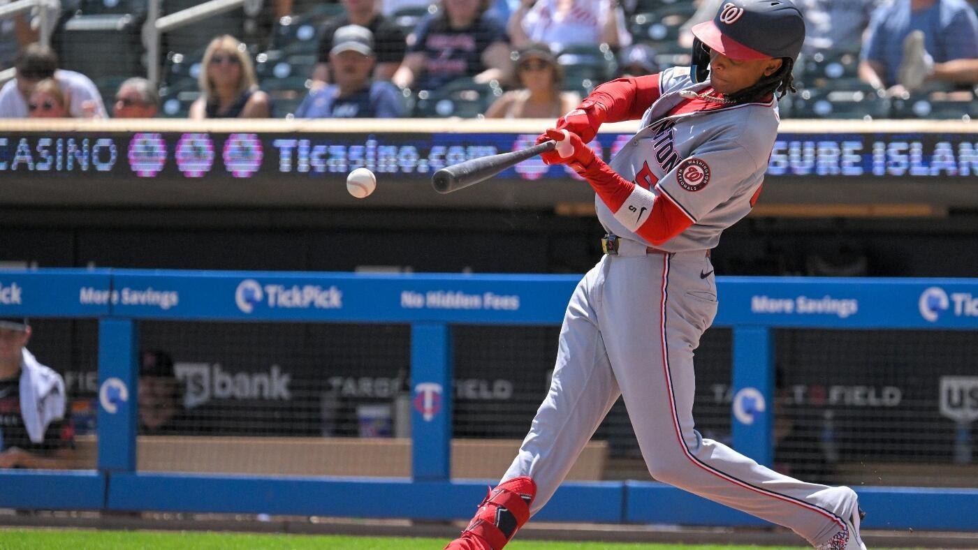 A baseball player in a gray Washington Nationals uniform swings a bat at an incoming ball during a game, with spectators visible in the stands behind him.