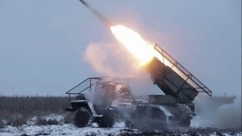 A truck-mounted multiple rocket launcher fires a rocket into the sky, producing a large burst of flame and smoke over a snowy field.