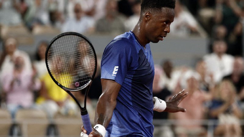 A male tennis player in a blue shirt holds a racket while looking downward, with a blurred crowd seated in the background.