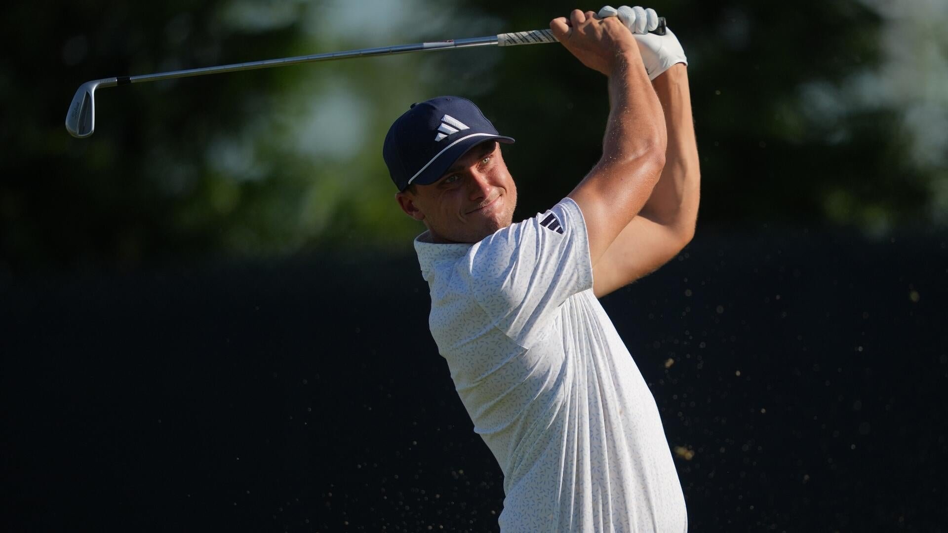 A golfer in a white patterned shirt and navy Adidas cap completes a swing, holding the club raised overhead against a dark green foliage background.