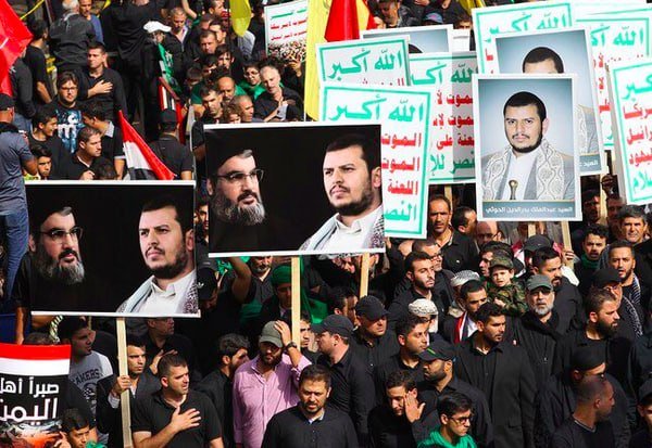 Large crowd of people in black clothing march while holding Arabic signs and large portraits of bearded men.