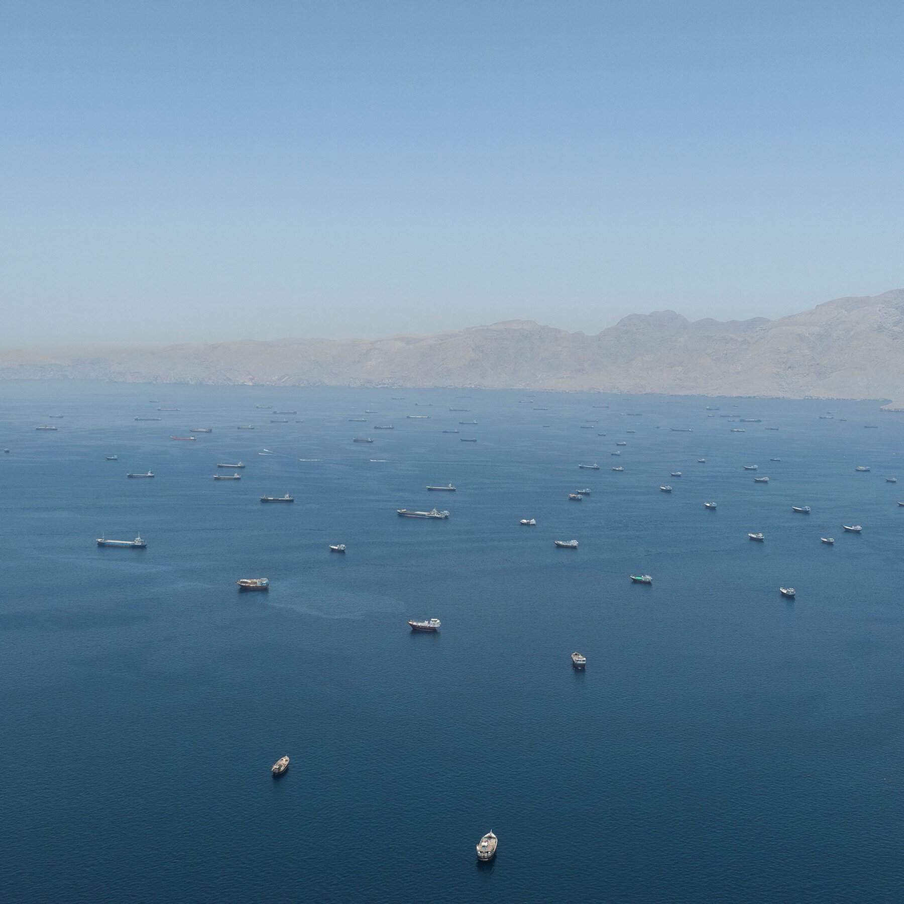 Dozens of cargo ships and vessels are anchored in a calm blue bay with arid, mountainous coastline visible in the background.