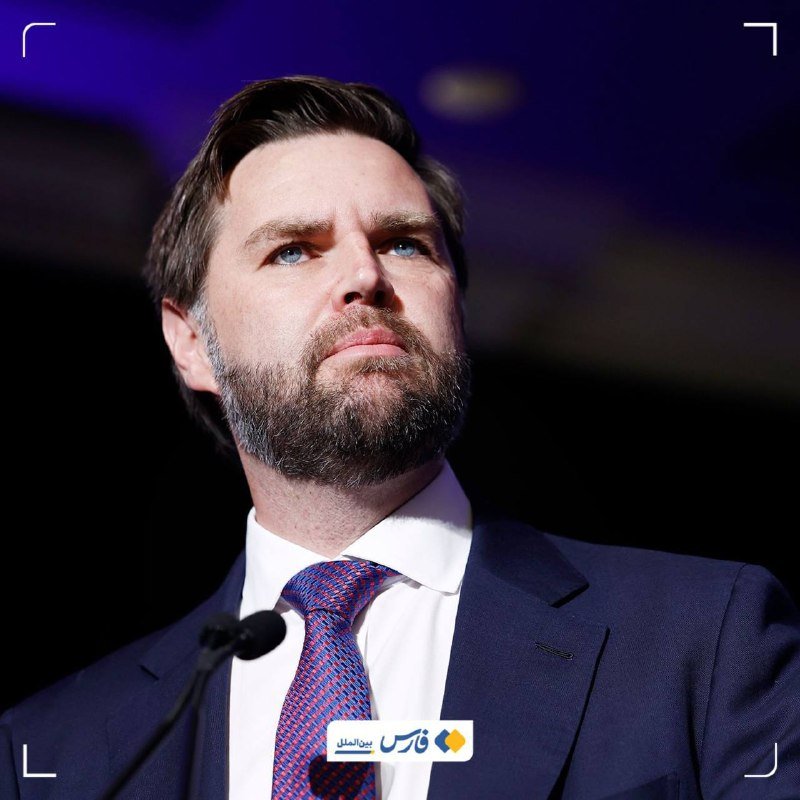 A bearded man in a navy suit and patterned tie stands at a microphone, gazing upward against a dark blue backdrop.